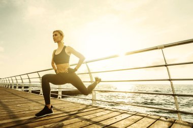 Deniz kenarındaki güvertede sağlıklı kadın eğitimi. Spor giyim sektöründe genç ve çekici bir kadın gün doğumunda plajda egzersiz yapmadan önce bacak esnetiyor. Isının.