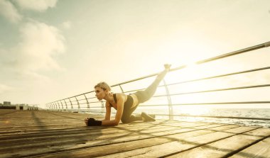 Deniz kenarındaki güvertede sağlıklı kadın eğitimi. Spor giyim sektöründe genç ve çekici bir kadın gün doğumunda plajda egzersiz yapmadan önce bacak esnetiyor. Isının.