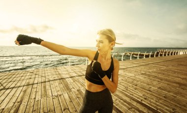 Sargı bezine sarılmış elleri olan genç bir kickboksör kadın gün doğumunda sahilde trenleri yumrukluyor. Sabah dövüşçüsü