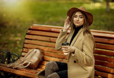 Sonbahar kıyafetleri içinde genç çekici bir kadın kenarda oturur ve şehir parkında kahve içer. Şık giyinmiş ve keçeli şapkalı bir kadın. Sonbahar zamanı