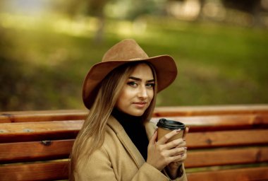 Sonbahar kıyafetleri içinde genç çekici bir kadın kenarda oturur ve şehir parkında kahve içer. Şık giyinmiş ve keçeli şapkalı bir kadın. Sonbahar zamanı