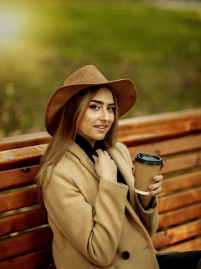 Sonbahar kıyafetleri içinde genç çekici bir kadın kenarda oturur ve şehir parkında kahve içer. Şık giyinmiş ve keçeli şapkalı bir kadın. Sonbahar zamanı