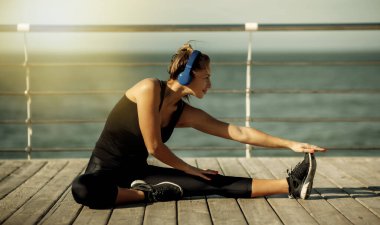 Spor kıyafetli zayıf bir kadın, güneşli bir günde bacaklarını esnetiyor ve plajda kulaklıkla müzik dinliyor.