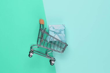 Mavi pastel arka planda fiyonklu alışveriş arabası ve hediye kutusu. Noel, doğum günü ya da düğün için kompozisyon. Üst görünüm