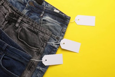 İçinde boş kağıt etiketler olan bir sürü değişik moda kot pantolonlar. Alışveriş, satış konsepti. Sarı arkaplan