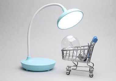 Masa LED lambası ve parlak ampul gri arka planda mini alışveriş arabasında.