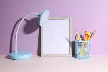 Masa lambası ve mor arkaplanda kırtasiye malzemesi olan boş çerçeve