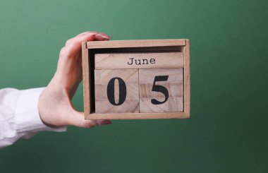 5 Haziran tarihli ahşap blok takvimini yeşil arka planda tutan bir el.