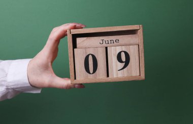 9 Haziran tarihli ahşap blok takvimini yeşil arka planda tutan bir el.