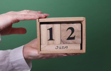 12 Haziran tarihli ahşap blok takvimini yeşil arka planda tutan bir el.