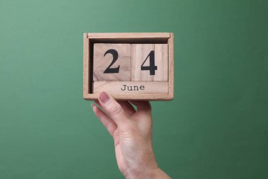 24 Haziran tarihli ahşap blok takvimini yeşil arka planda tutan bir el.