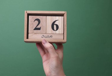26 Haziran tarihli ahşap blok takvimini yeşil arka planda tutan bir el.