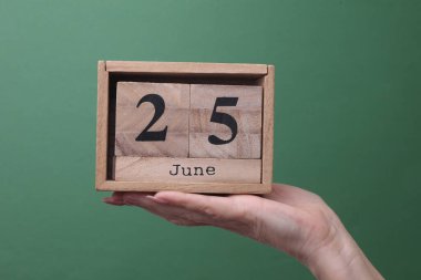 25 Haziran tarihli ahşap blok takvimini yeşil arka planda tutan bir el.