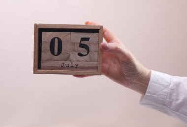 Kadın El, tahta blok takvimi tutuyor. Bej arka planda 5 Temmuz tarihi var.