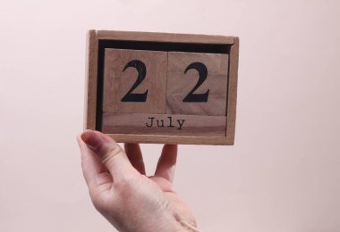 Kadın El, tahta blok takvimi tutuyor. Bej arka planda 22 Temmuz tarihi var.