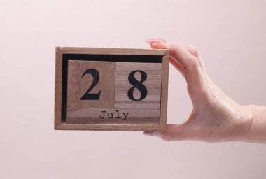 Bayan El, tahta blok takvimi tutuyor. Bej arka planda 28 Temmuz tarihi var.