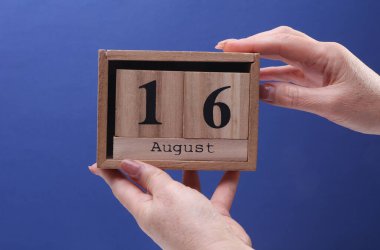 16 Ağustos tarihli ahşap blok takvimi tutan kadın el mavi arka planda
