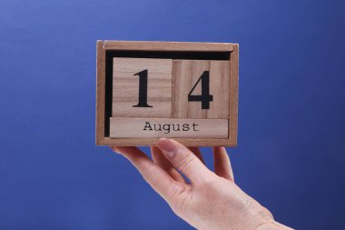 14 Ağustos tarihli ahşap blok takvimi tutan kadın el mavi arka planda.