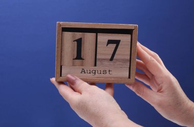 17 Ağustos tarihli ahşap blok takvimi tutan kadın el mavi arka planda.