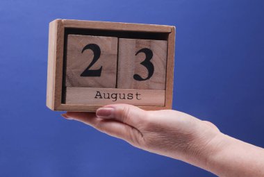23 Ağustos tarihli ahşap blok takvimi tutan kadın el mavi arka planda.
