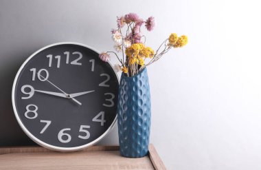 Kuru çiçekli vazo, ahşap tezgahta saat, gri arka plan.