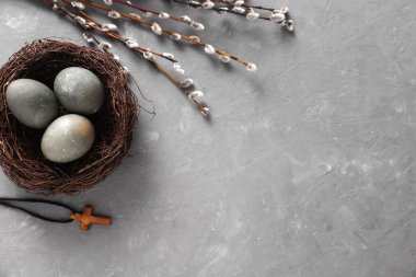Easter eggs in nest with Christian cross and pussy willow branches on grey concrete background. Top view
