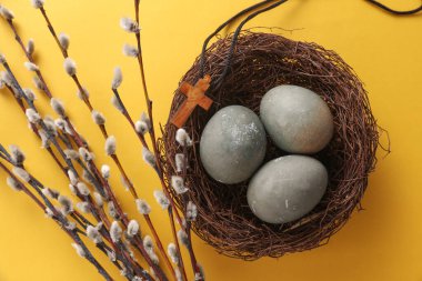 Easter eggs in nest with Christian cross and pussy willow branches on yellow background. Top view