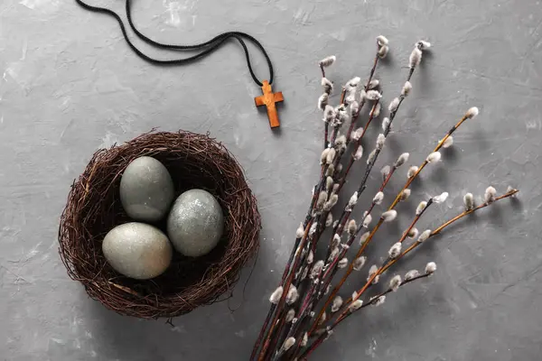 Easter eggs in nest with Christian cross and pussy willow branches on grey concrete background. Top view