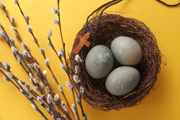 Easter eggs in nest with Christian cross and pussy willow branches on yellow background. Top view
