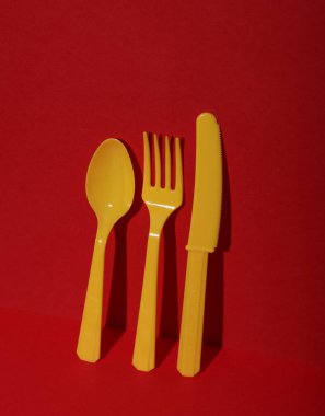 Sarı plastik kaşık, kırmızı arka planda bıçak ve çatal, gölgeli. Minimalizm. Yaratıcı düzen. Yiyecek konsepti