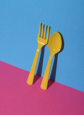 Sarı plastik kaşık ve çatal pembe mavi arka planda, gölgeli. Minimalizm. Yaratıcı düzen. Yiyecek konsepti