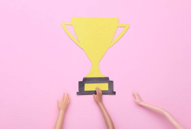 Doll 'un elleri pembe arka planda altın bir kupa tutuyor. Yarışma, zafer.