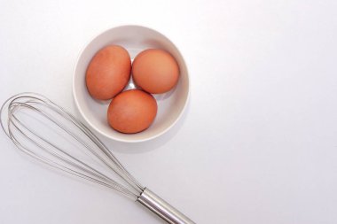 Top view of chickken eggs in a bowl and egg whisks on white background. Copy space.