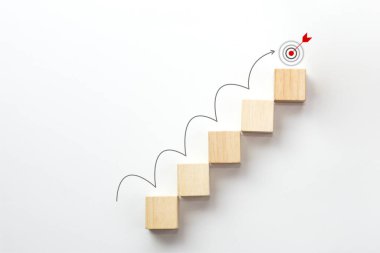 Tahta blok beyaz zemin üzerinde ok yükselen basamak merdiven olarak istifleniyor ve hedef simge üzerinde. İş büyümesi, başarı, strateji kavramı.