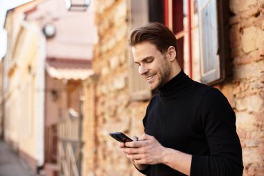 Yakışıklı bir adam akıllı telefonuyla meşgul olurken büyüleyici bir sokakta gülümsüyor..