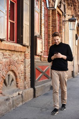Şık bir adam telefonunu kullanırken çekici bir kaldırım taşı caddesinde rahatça dolaşır..
