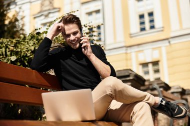 Şık bir adam bankta oturur, telefonda konuşur ve dizüstü bilgisayarı üzerinde çalışırken gülümser..