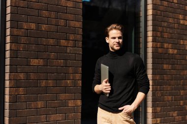 Şık bir adam elinde bir dizüstü bilgisayarla, kendine güveni ve cazibesi ile canlı bir şehirde gezinir..