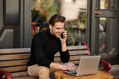 Şık bir adam bir kafede dizüstü bilgisayarını yazarken canlı bir telefon görüşmesi yapar..