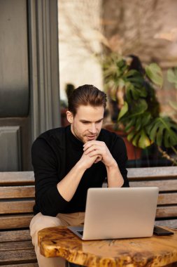 Düşünceli bir ifadeye sahip genç bir adam büyüleyici bir kafede laptopuna dikkatle odaklanır..