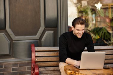 Yakışıklı bir adam dışarıda dizüstü bilgisayarı üzerinde çalışırken ahşap bir masaya gülümsüyor..