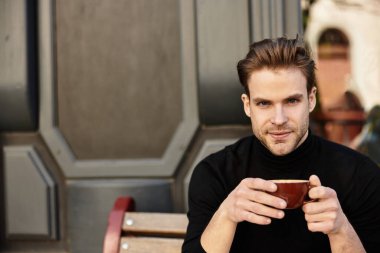 Çekici bir genç adam bir bankta oturuyor, canlı bir şehir ortamında sıcak bir içeceğin tadını çıkarıyor..