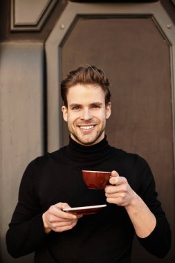 Şık giyinmiş yakışıklı bir adam sokakta yürüyor, elinde kahve tutarken gülümsüyor..