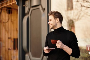 Neşeli gülüşü olan bir adam elinde fincan ve fincan tabağı tutarken tuhaf bir açık hava ortamında yürüyor..