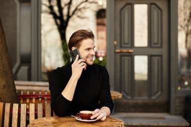 Yakışıklı adam dışarıda canlı bir şehir kafesinde yürüyüp, telefonla konuşuyor ve kahvenin tadını çıkarıyor..
