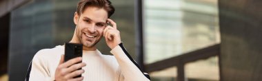 Yakışıklı bir adam akıllı telefonuyla konuşurken gülümseyen bir şekilde şehir ortamında gezinir..