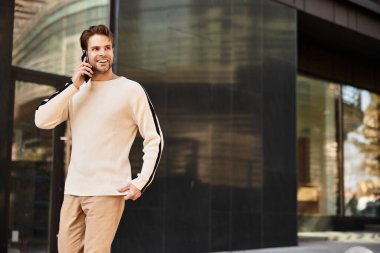 Parlak giyimli beyefendi güvenli bir şekilde yürüyor, modern bir şehir binasının yakınında telefonla konuşuyor..