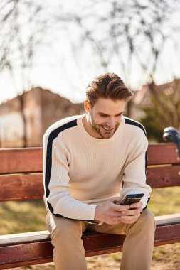 Çekici bir adam, canlı bir parkta, açık gökyüzünün altında telefonunu kullanarak bir bankta gülümsüyor..