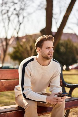 Yakışıklı bir adam elinde telefonla güneşli bir parkta geziniyor, gülümsüyor ve hayatı yansıtıyor..