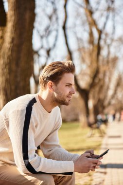 Genç, yakışıklı bir adam, sakin bir parkta akıllı telefonuna göz atmak için yürüyüşüne ara veriyor..